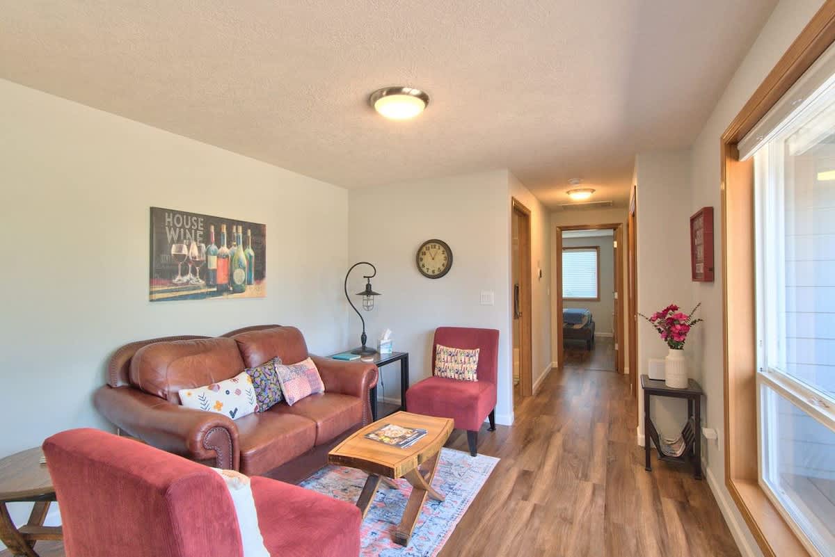 Cozy living room featuring a brown leather sofa, a red accent chair, a rustic wooden coffee table, and light wood laminate flooring, with a hallway leading to other rooms.