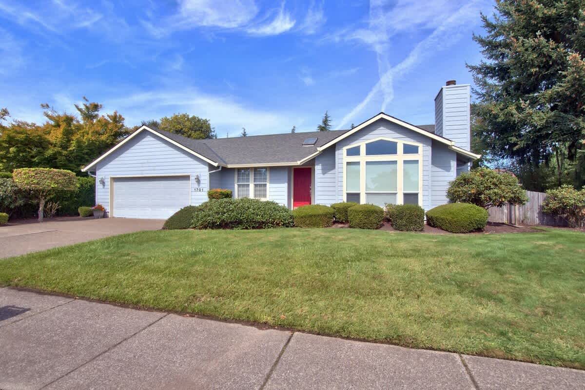 A charming single-level light blue house with a red front door, a two-car garage, and a well-maintained green lawn. The house features a large bay window and a chimney, surrounded by mature landscaping.