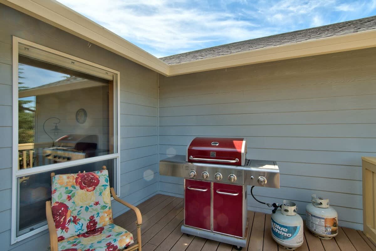 A red and stainless steel gas grill with two propane tanks sits on the wooden deck next to a large window, ready for outdoor cooking.