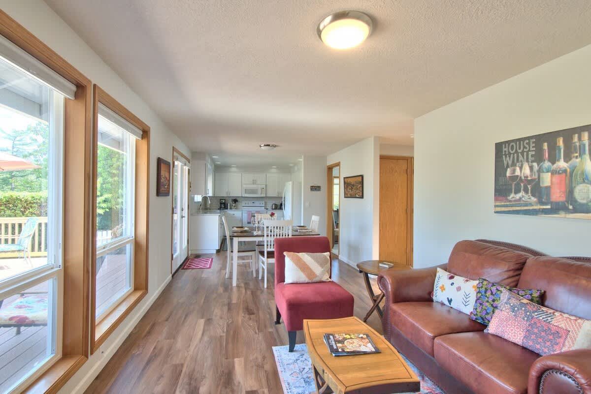 Open-concept living space featuring a brown leather sofa with colorful throw pillows, a red accent chair, and a wooden coffee table, all on a light wood floor, leading to a dining area and kitchen.