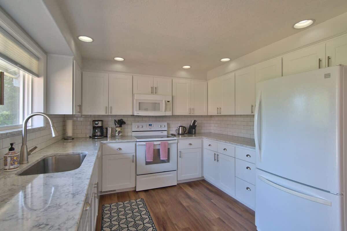 U-shaped kitchen with white shaker cabinets, light granite countertops, and white appliances including a refrigerator, range, and dishwasher, all on a light wood floor.