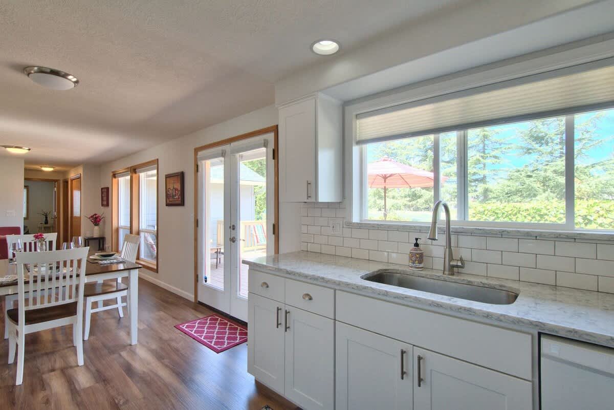 Modern kitchen with white shaker cabinets, light granite countertops, a stainless steel sink under a large window, and subway tile backsplash, with a view of the dining area and outdoor deck.