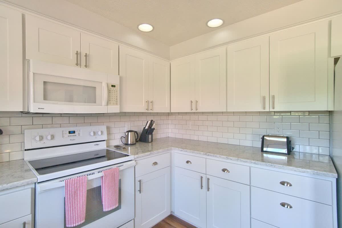 Close-up of a modern white kitchen corner with shaker cabinets, light granite countertops, a white electric range, and an over-the-range microwave, set against a white subway tile backsplash.