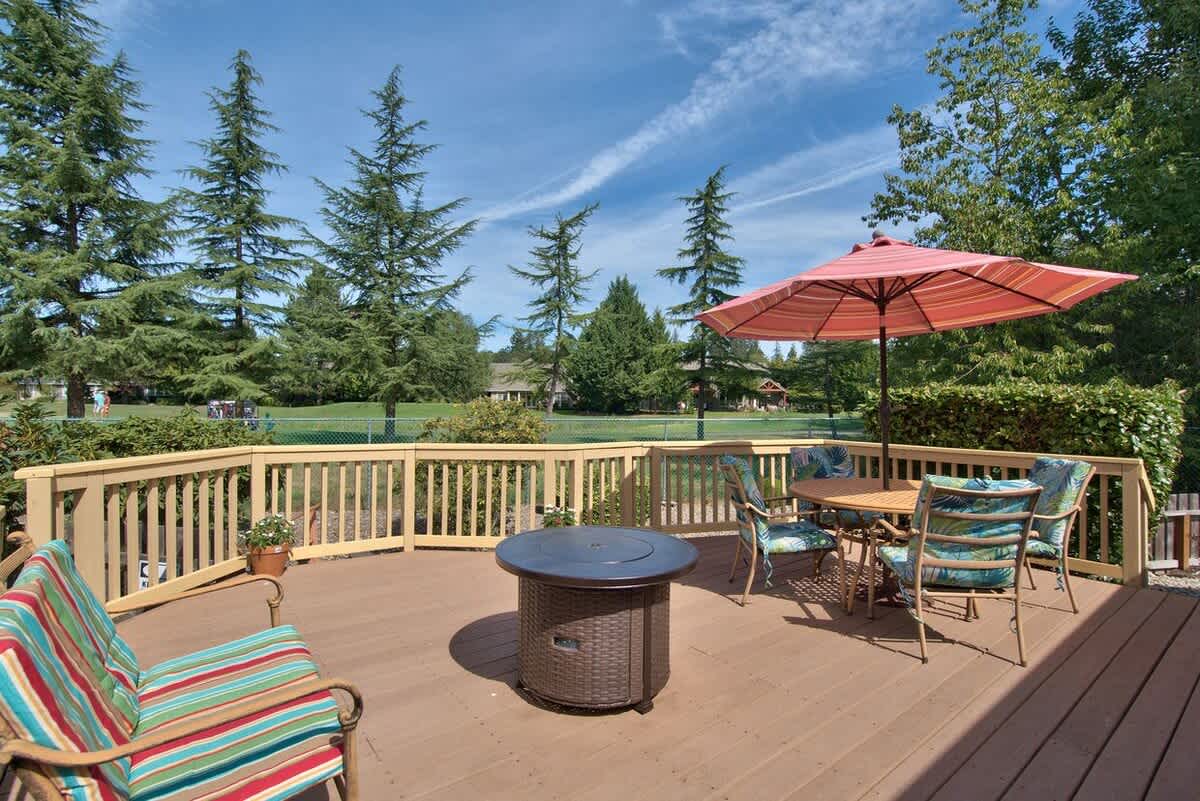 Spacious outdoor deck furnished with a dining table under a red umbrella, a fire pit table, and various seating options, overlooking a lush green golf course.