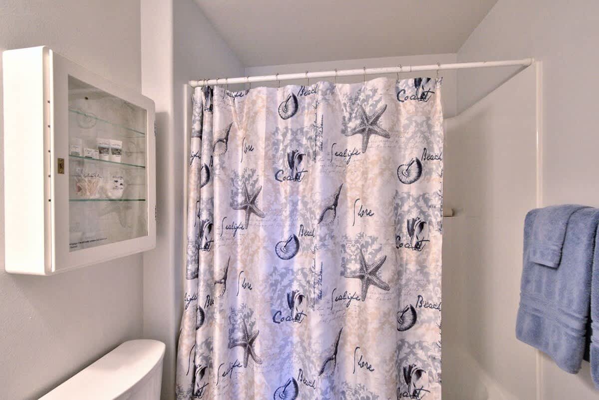 Close-up view of the bathroom's shower area, featuring a white shower base and a decorative shower curtain with starfish and seashell patterns. A white medicine cabinet is visible above the toilet.