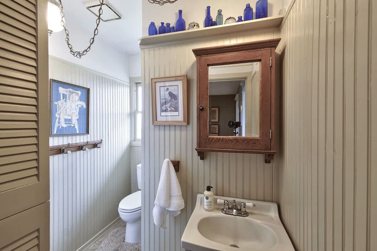 Bathroom with light-colored beadboard walls, a pedestal sink, and a toilet. A wooden medicine cabinet with a mirror hangs above the sink, and blue bottles are displayed on a shelf.