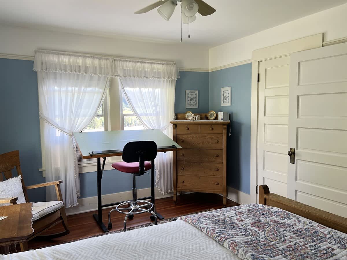 A bright bedroom with light blue walls, a wooden dresser, a drafting table, and a window with white sheer curtains, offering a view outside.