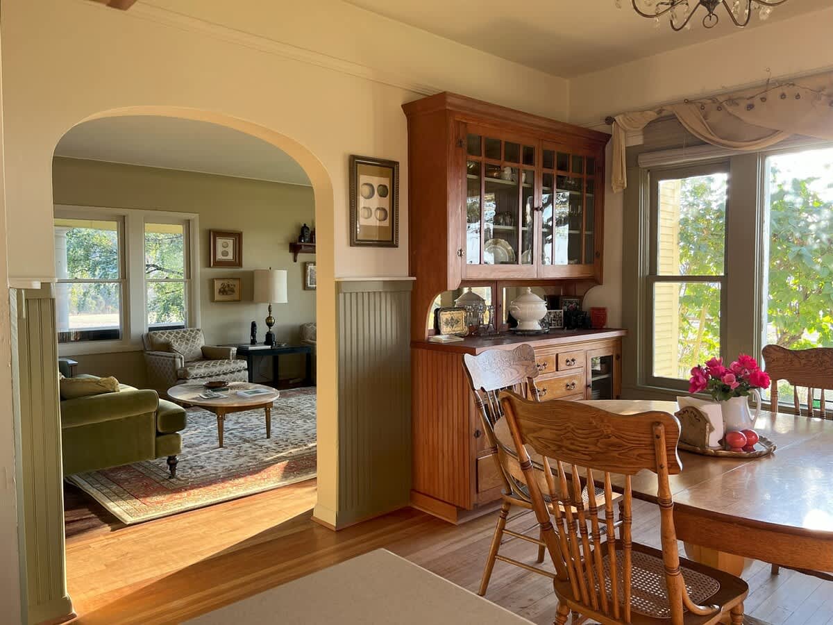 View into the dining room through an arched doorway, revealing a wooden dining table, a large wooden hutch, and a window offering a glimpse of the outdoor landscape.