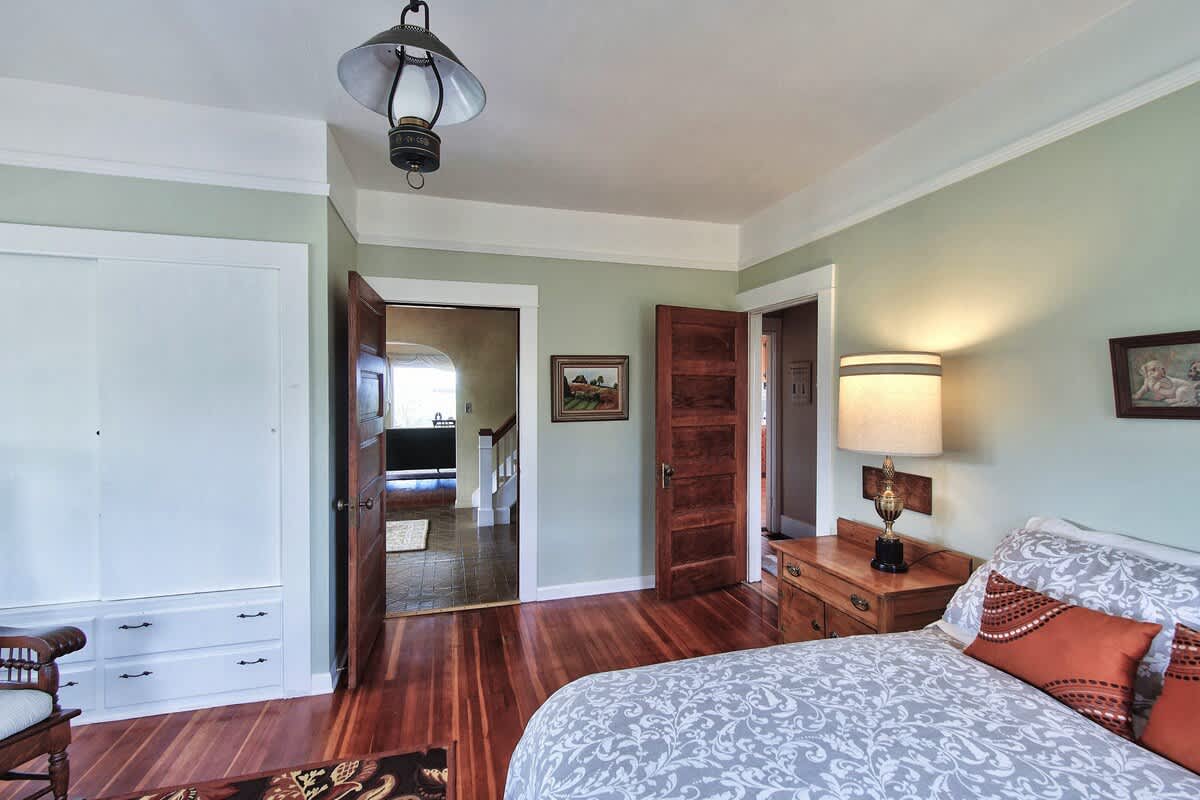 Another perspective of the bedroom, highlighting a built-in closet with sliding white doors and drawers. A doorway leads to a hallway with stairs, and the room maintains its light green walls and hardwood floors.