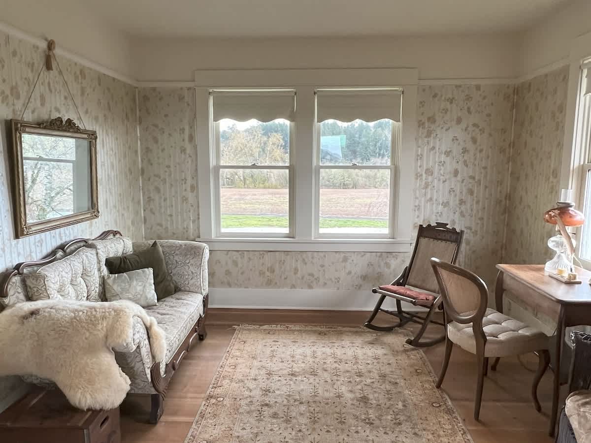 Inviting living space with patterned wallpaper, light wood floors, and a vintage-style sofa with a sheepskin throw. A rocking chair, a small desk, and large windows offering outdoor views complete the cozy ambiance.