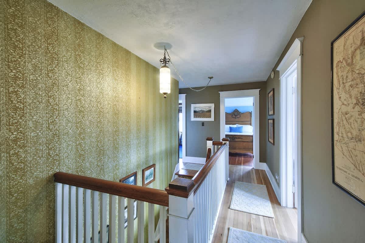 A brightly lit hallway with a white banister and wooden handrail, featuring green patterned wallpaper and hardwood floors, leading to other rooms upstairs.