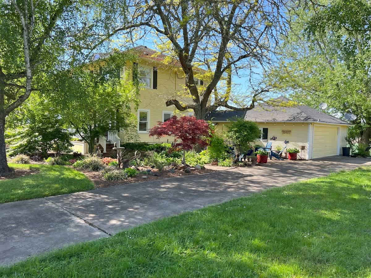 A charming yellow two-story farmhouse with dark trim is nestled among lush green trees and landscaping, featuring a paved driveway leading to a garage, all under a bright sunny sky.