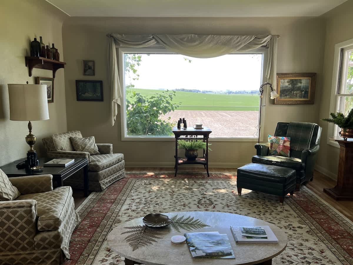 Cozy living room with a large picture window offering expansive views of rolling fields, featuring a patterned area rug, two upholstered armchairs, and a central coffee table.