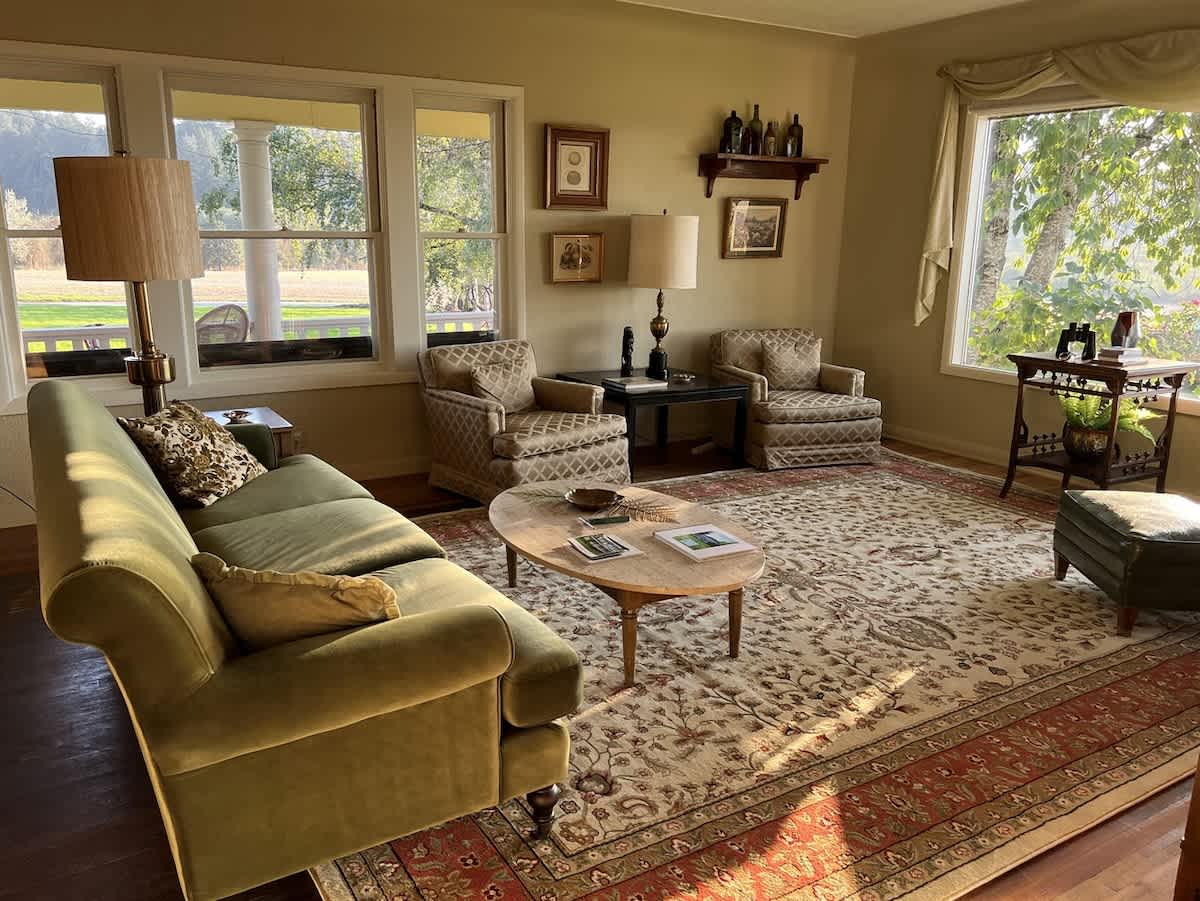 This inviting living room is furnished with a comfortable green sofa, two elegant patterned armchairs, and a large decorative area rug, all illuminated by abundant natural light from several windows.