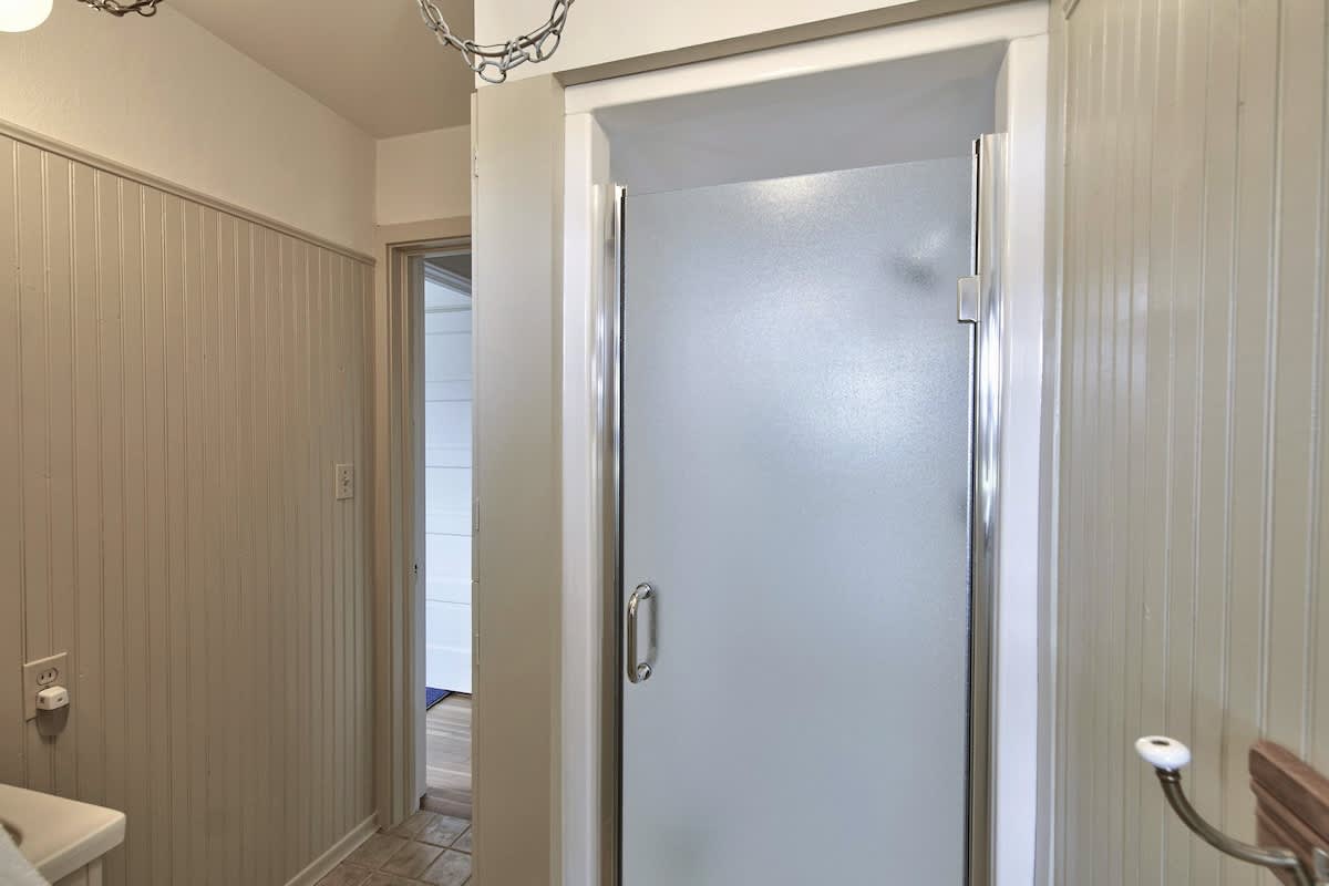 Bathroom with light-colored beadboard walls and a frosted glass shower enclosure. A doorway leads to another room, and the floor appears to be tiled.