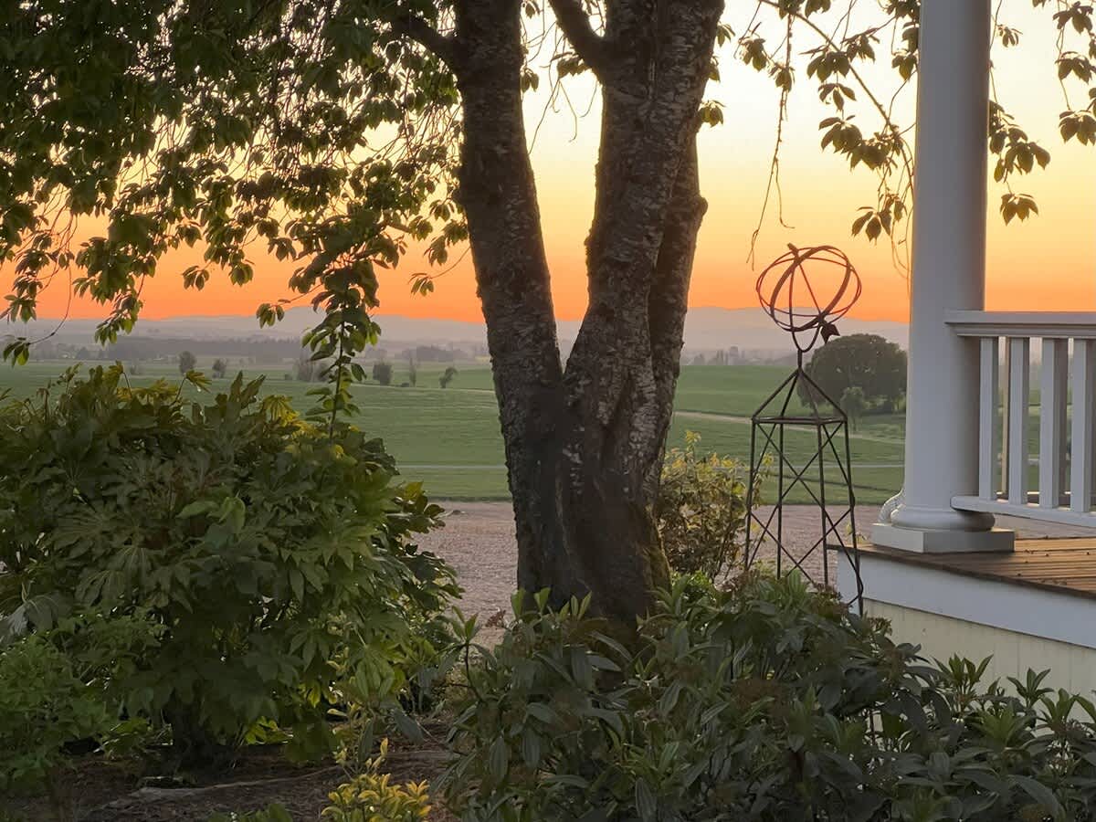 A close-up view from the porch reveals a vibrant sunset over rolling fields and distant mountains, with an ornate metal garden sphere adding a decorative touch near a white column and lush green bushes.