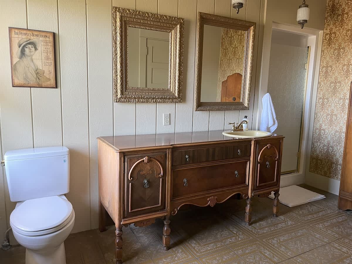 A vintage-style bathroom with a white toilet, a dark wood vanity featuring a cream sink, and two ornate gold-framed mirrors, set against vertical wood paneling and patterned floor tiles.