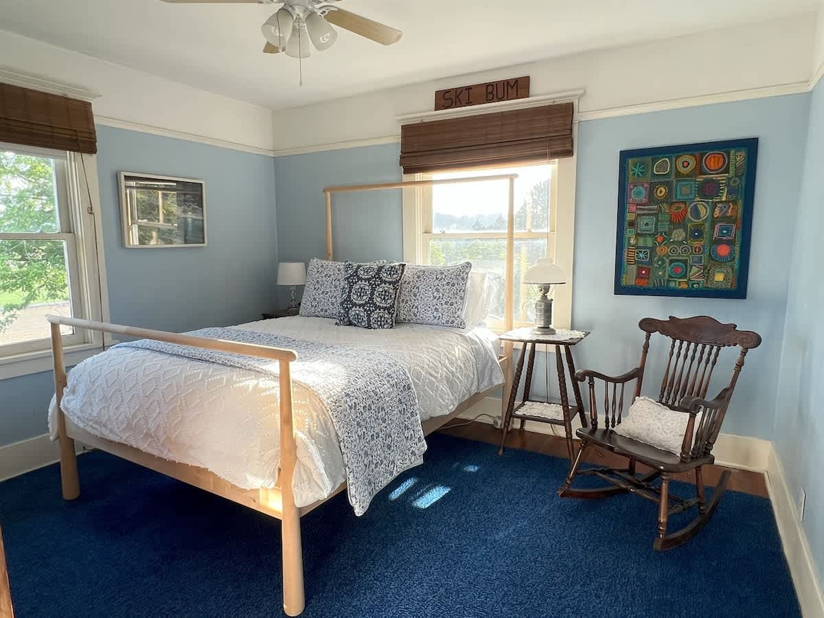 Cozy bedroom with light blue walls, a light wood bed frame, and blue patterned bedding. A blue carpet covers the floor, and a rocking chair sits beside a window with a view. A "SKI BUM" sign is above the window.