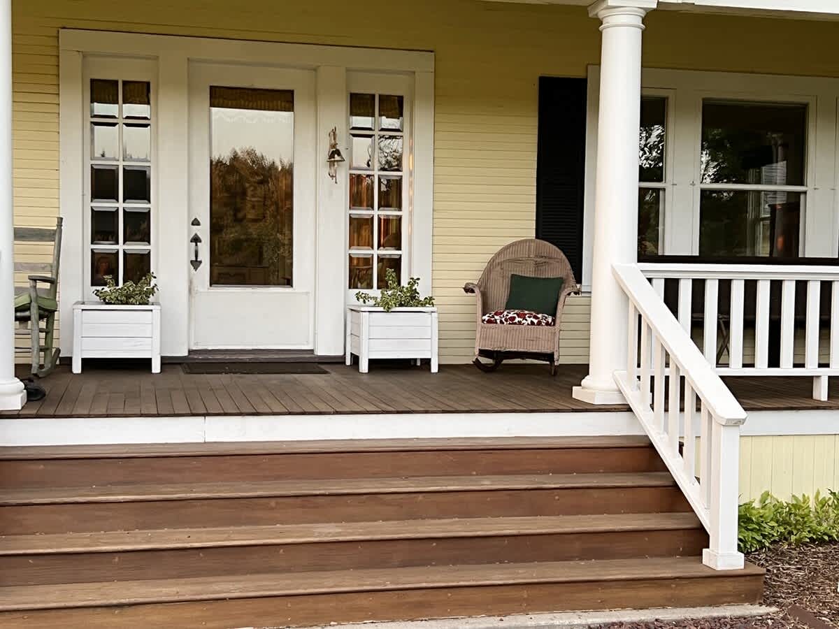 A close-up of the inviting front porch entrance, showcasing white columns, sturdy wooden steps, and a wicker rocking chair with a green cushion, flanked by white planters and a glass-paned front door.
