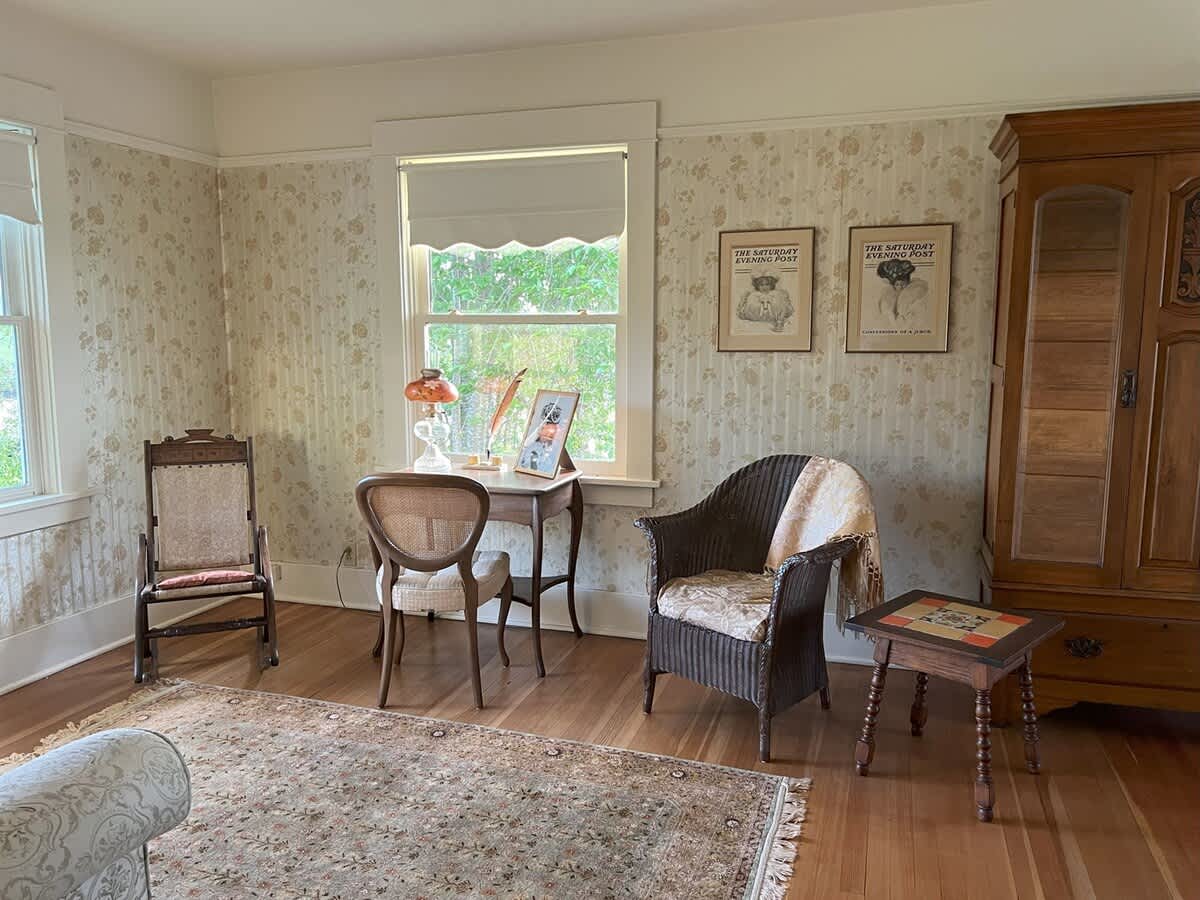 A vintage-style living room with floral wallpaper, a wooden desk and lamp, a wicker armchair, and a patterned area rug on hardwood floors, complemented by a large wooden armoire.