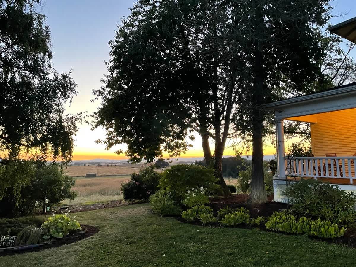 The well-manicured lawn leads towards the illuminated porch of the yellow house, framed by large trees, with a breathtaking sunset painting the distant sky in warm orange and yellow hues over vast fields.