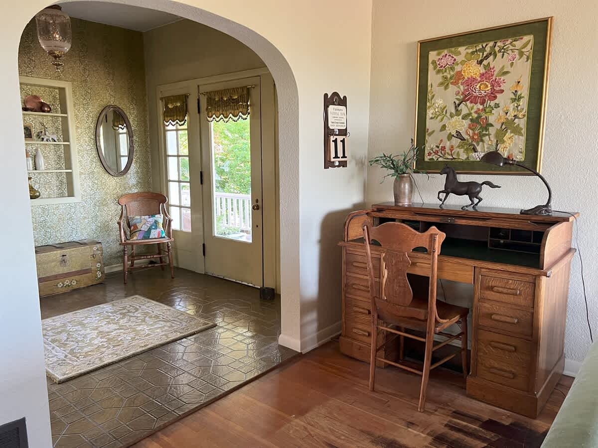 A dedicated office nook, adjacent to the foyer, features a traditional wooden roll-top desk with a matching chair, a vibrant floral artwork, and a window providing natural light.