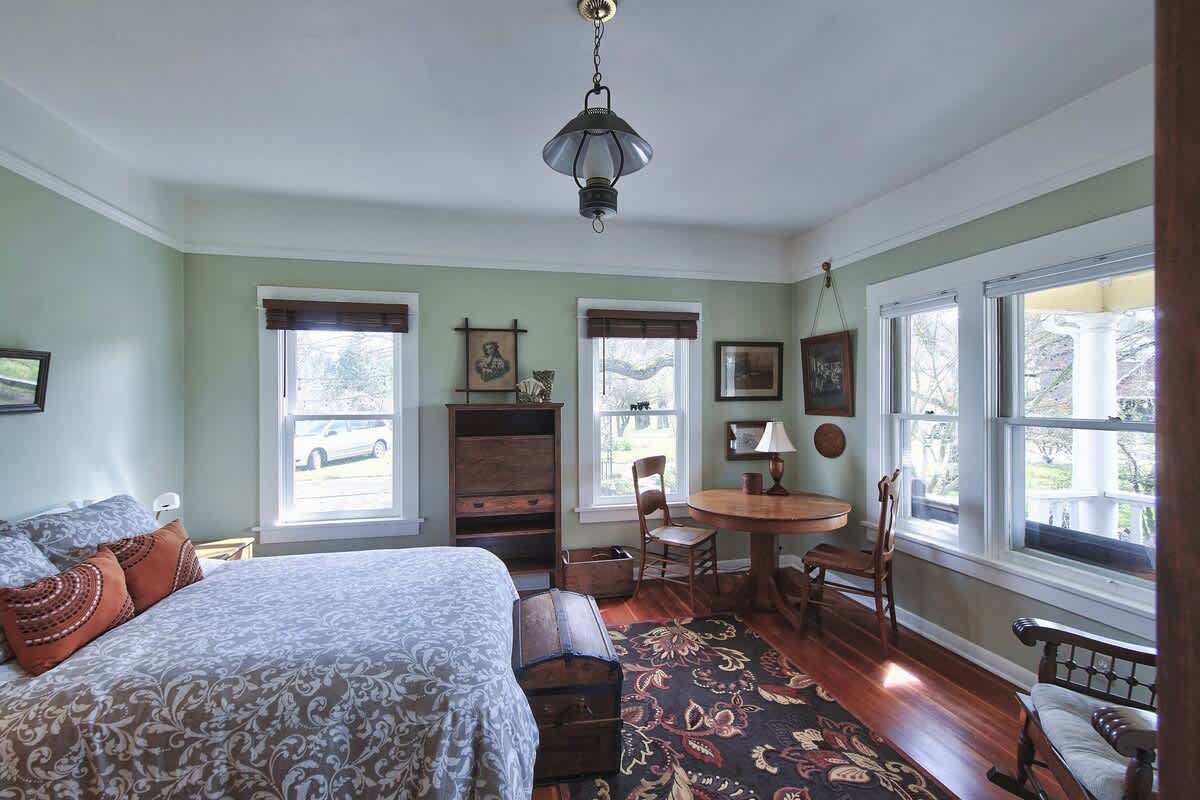 Cozy bedroom with light green walls, hardwood floors, and a bed with a patterned white and grey duvet. A round wooden table with two chairs and a rocking chair are positioned near windows offering an outdoor view.