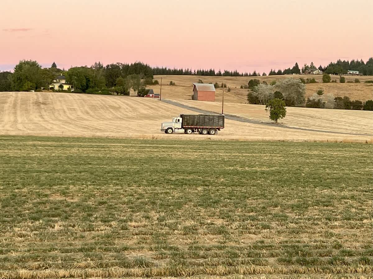 A large white semi-truck with a dark cargo bed drives on a dirt road through golden fields, with a red barn and distant trees under a soft pink and orange sunset sky.
