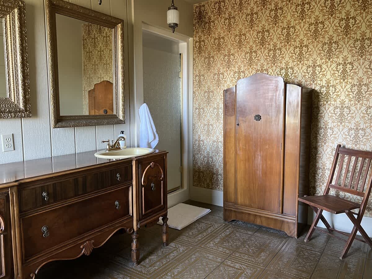 A vintage bathroom featuring a dark wood vanity with a cream sink, ornate mirrors, a tall wooden armoire, and a shower entrance, with patterned wallpaper and floor tiles.