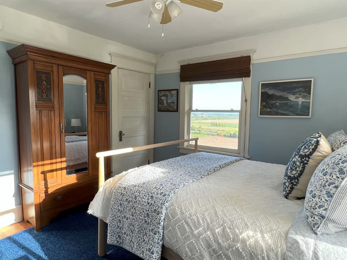 Light blue bedroom featuring a light wood bed frame with blue and white bedding, a large wooden armoire with a mirror, and a window overlooking green fields. Sunlight streams into the room, highlighting the blue carpet.