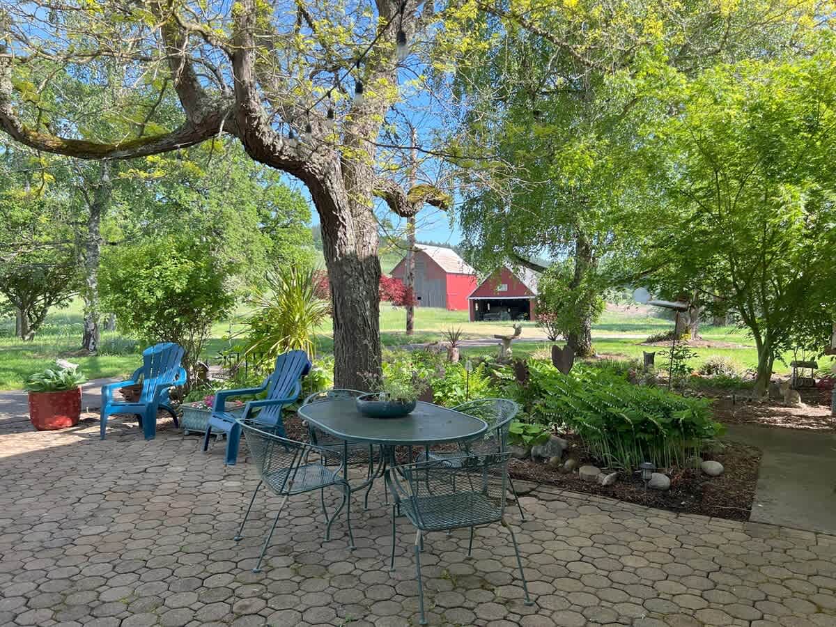 The stone-paved patio, shaded by a large tree, features a green metal dining set and blue Adirondack chairs, offering a picturesque view of a red barn and expansive green fields in the distance under a clear sky.