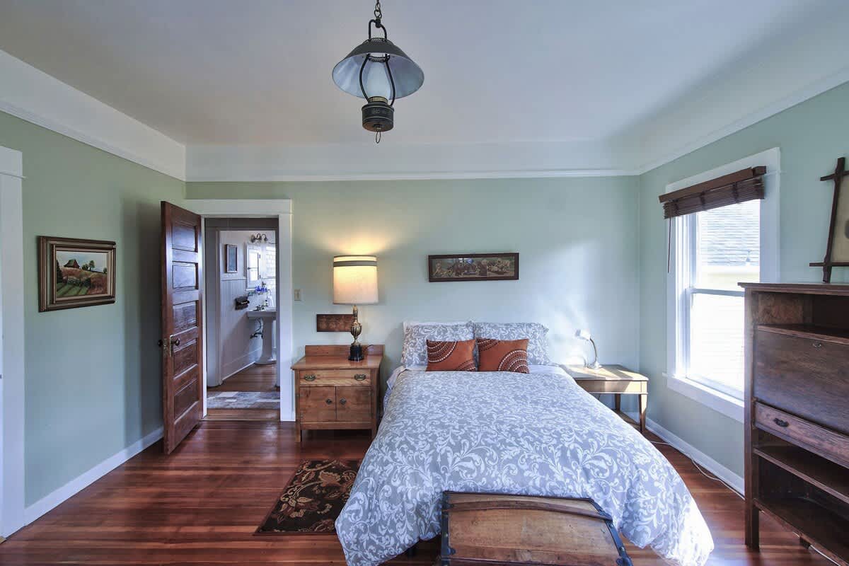 View of the same bedroom from a different angle, showing a wooden dresser and a bedside table with a lamp. A doorway leads to a bathroom, and another door opens to a hallway.