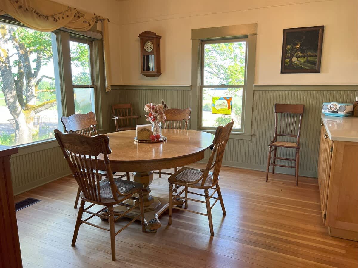 Inviting dining room with a round wooden table, various wooden chairs, and green beadboard wainscoting, featuring a wall-mounted clock and a window with outdoor views.