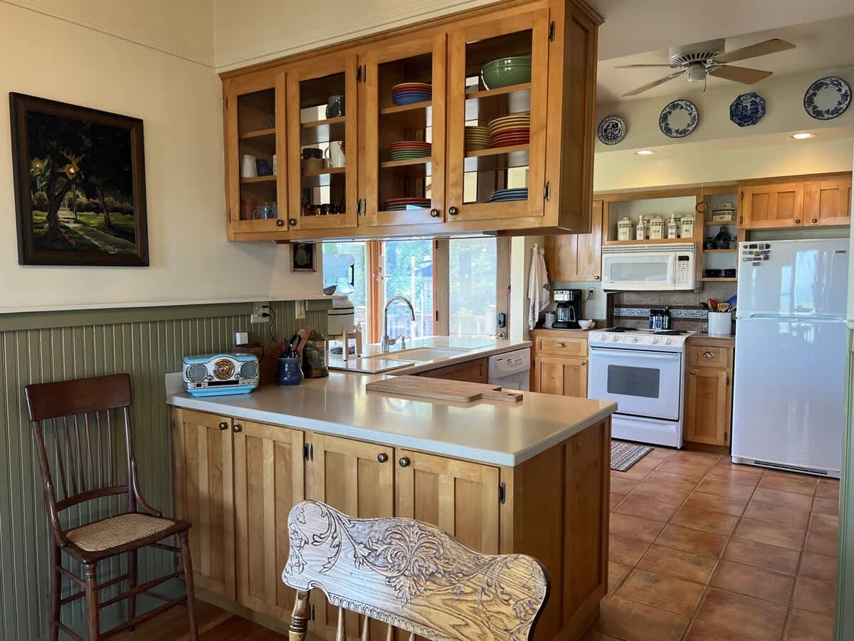 Traditional kitchen with light wood cabinetry, a central island with a light countertop, and a view towards the dining area, featuring a retro radio and tiled floors.