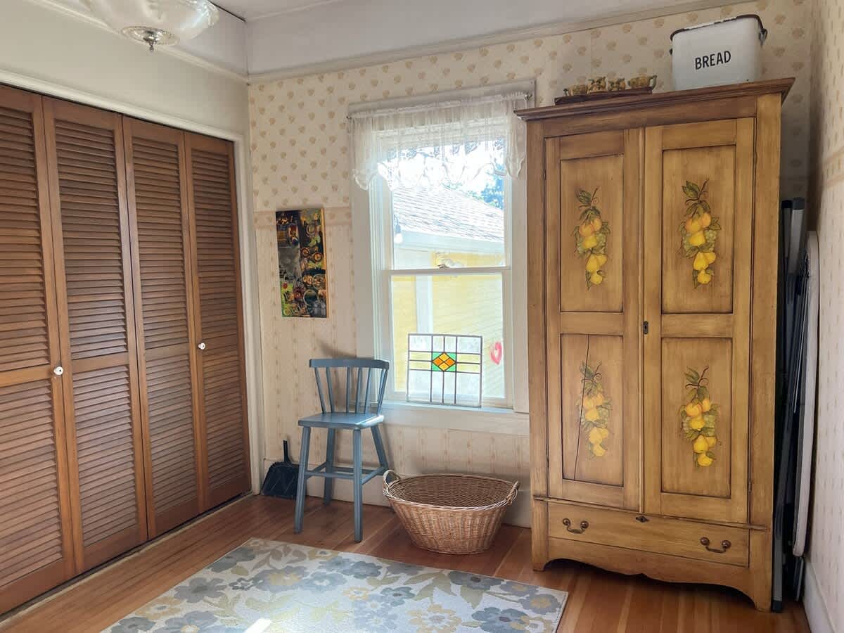Room with light patterned wallpaper, louvered wooden closet doors, and a large painted wooden armoire with lemon motifs. A window with a stained glass panel and a small blue chair are also present.