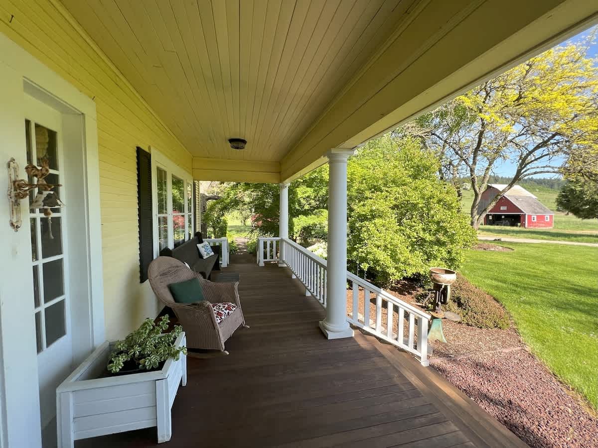 The cozy porch offers a wicker rocking chair and a swing, providing a relaxing spot with a clear view of a red barn and lush green trees in the distance, embodying a serene farm setting.