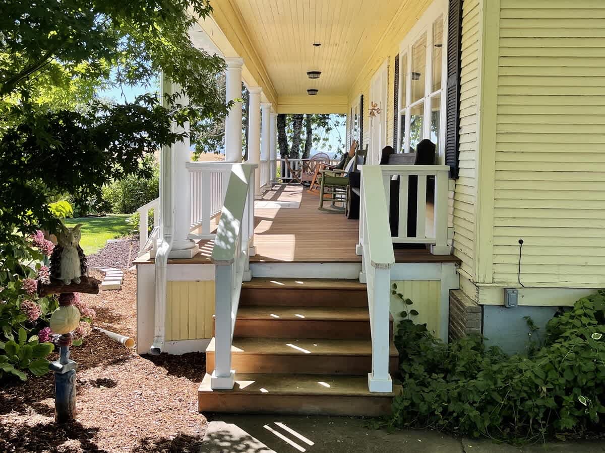 A side view of the expansive wrap-around porch, featuring white balusters, wooden flooring, and multiple seating areas, extending along the yellow-sided house under the shade of mature trees.