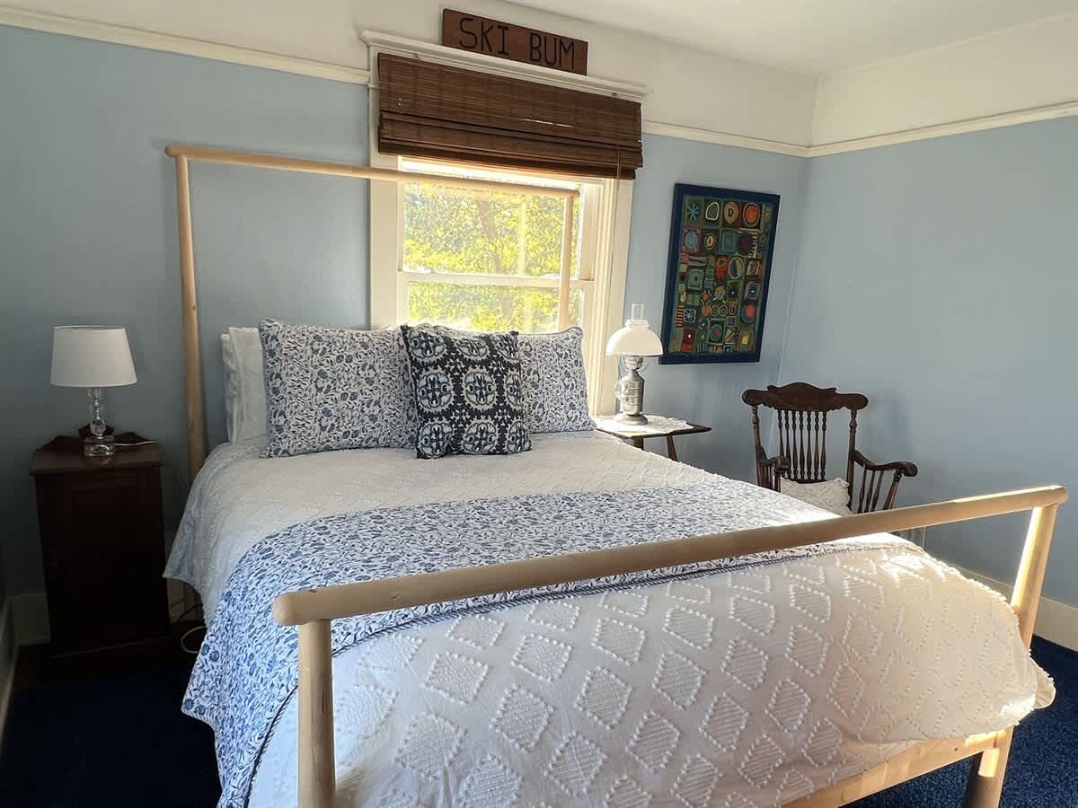 Bright bedroom with light blue walls, a light wood bed frame, and blue and white patterned bedding. A blue carpet covers the floor, and a bedside table with a lamp is next to the bed. A "SKI BUM" sign is above the window.