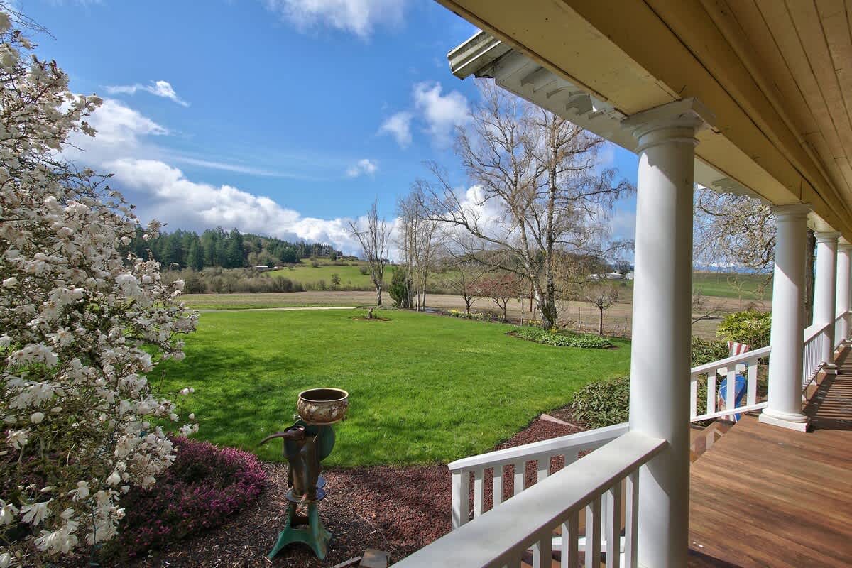 A picturesque view from the porch overlooks a sprawling green lawn and distant rolling hills, with white flowering bushes in the foreground and a bright blue sky with scattered clouds.