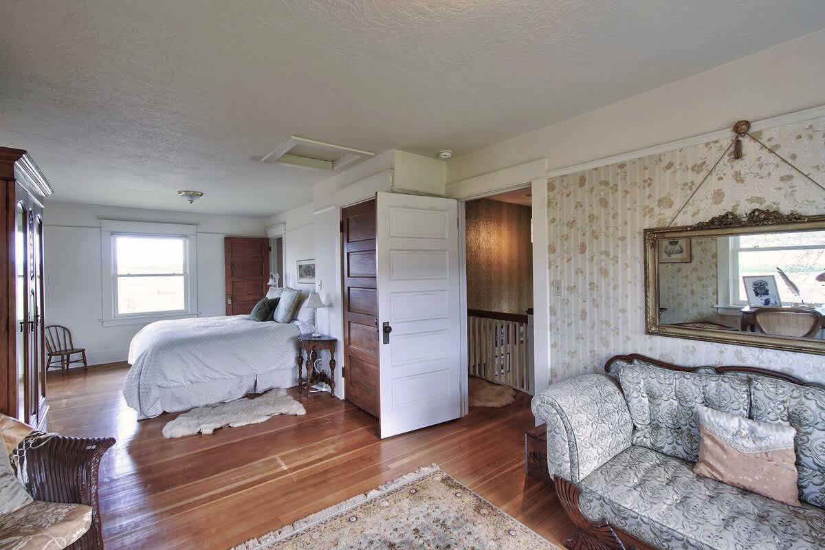 This bright bedroom features polished hardwood floors, a comfortable bed with white linens, a patterned chaise lounge, and a large ornate gold-framed mirror reflecting the room's classic decor.
