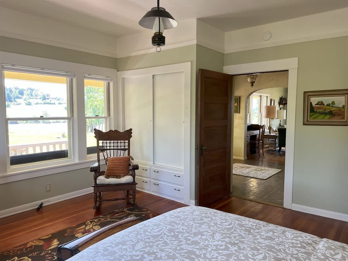 This image shows the bedroom with a large window offering a view of the outdoors. A rocking chair is placed near the window, and a built-in closet with drawers is visible.