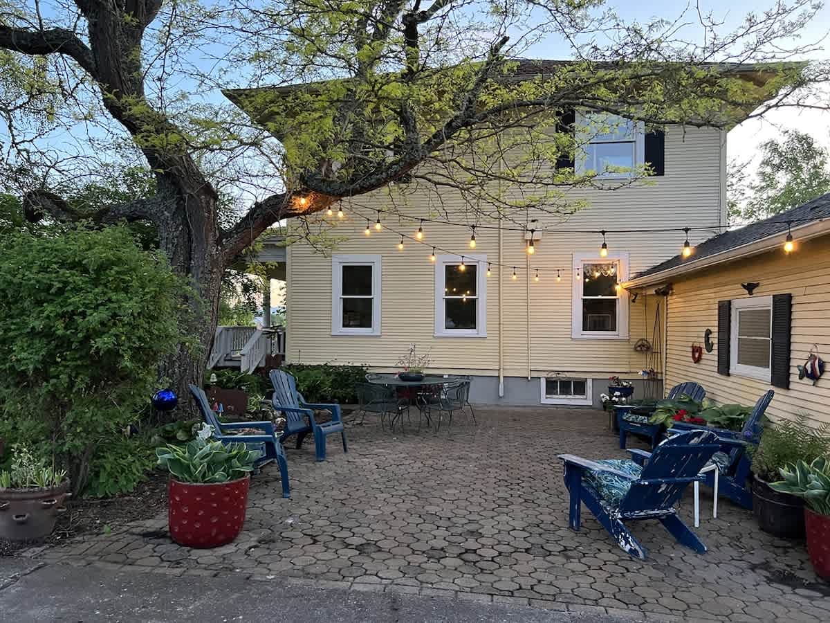 A spacious paved patio with interlocking pavers is set behind the yellow house, furnished with blue Adirondack chairs and a metal dining set, illuminated by string lights hanging overhead, creating a cozy evening ambiance.