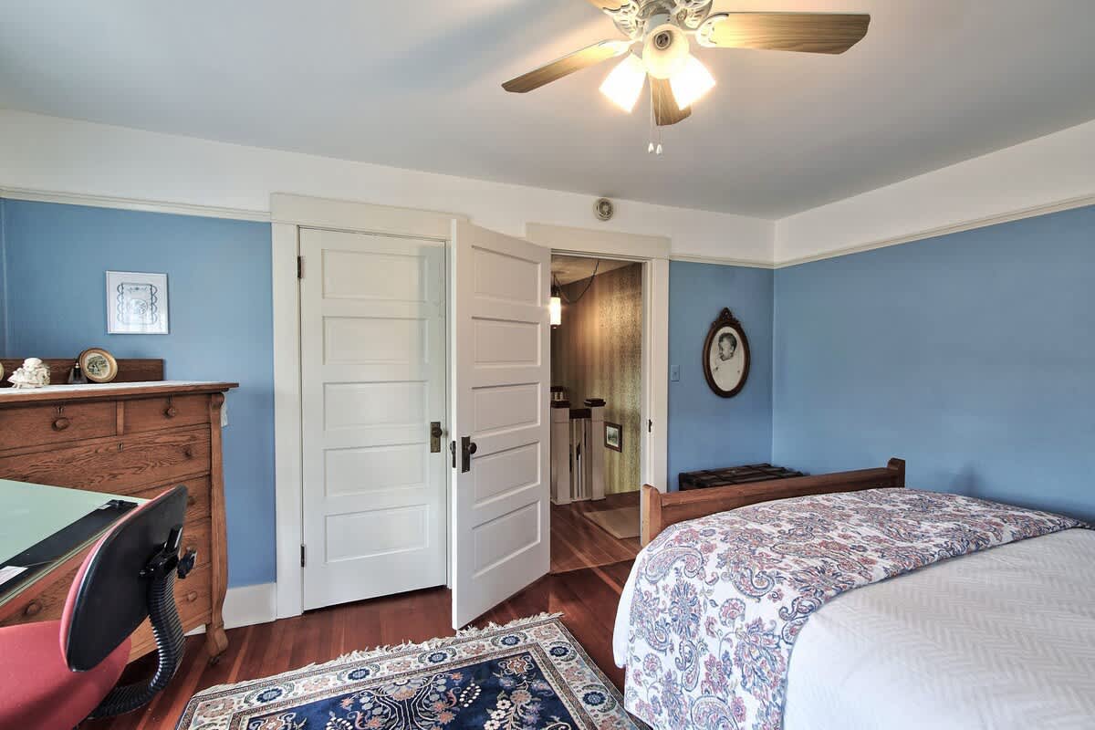 A cozy bedroom with light blue walls and white trim, featuring a wooden bed, a drafting table, and a dresser, with an open doorway leading to a hallway.