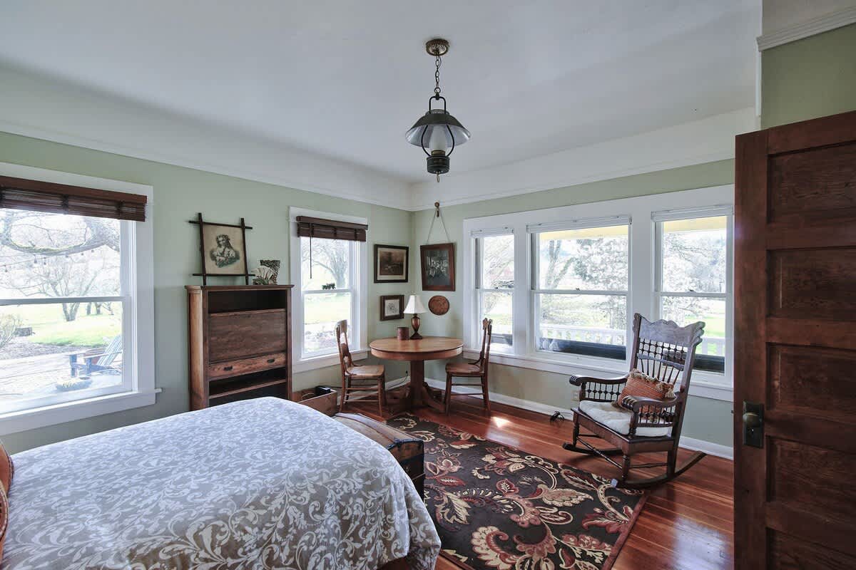 Wide shot of the bedroom, showcasing the bed, a small dining area with a round table and chairs, and a rocking chair. Multiple windows provide ample natural light and views of the surrounding landscape.