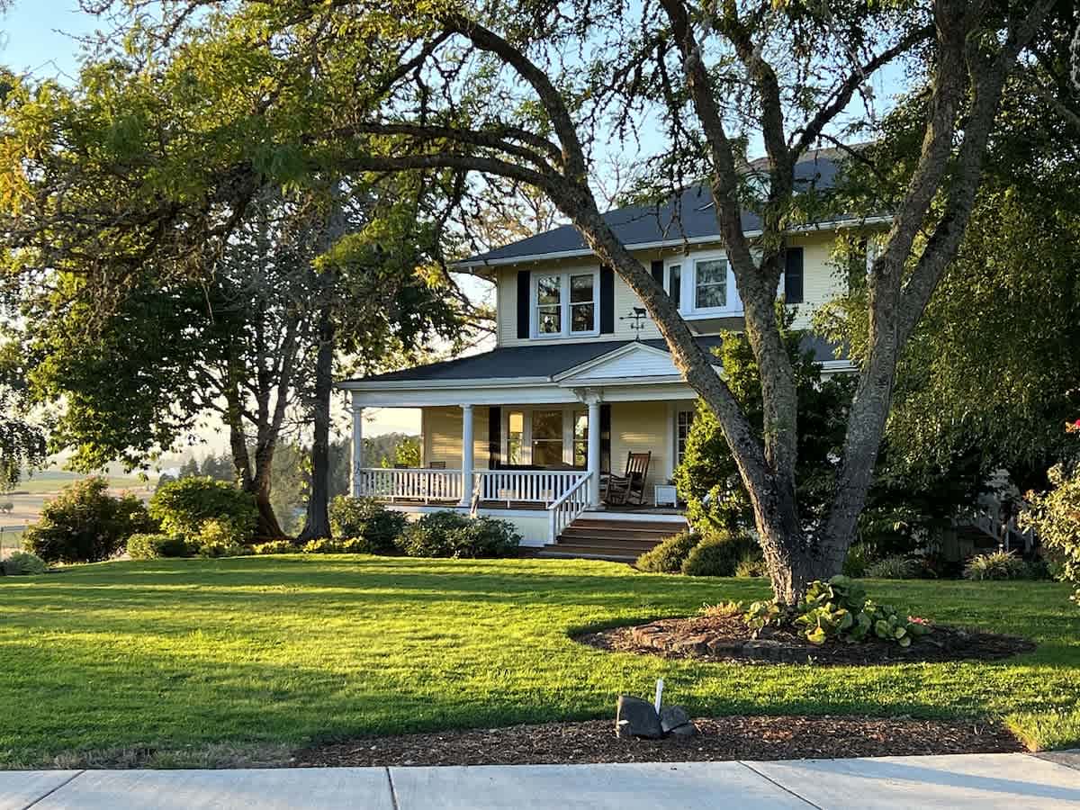 The grand yellow 1911 farmhouse, featuring a wrap-around porch with white railings, is nestled among mature trees and a manicured green lawn, bathed in warm sunlight.