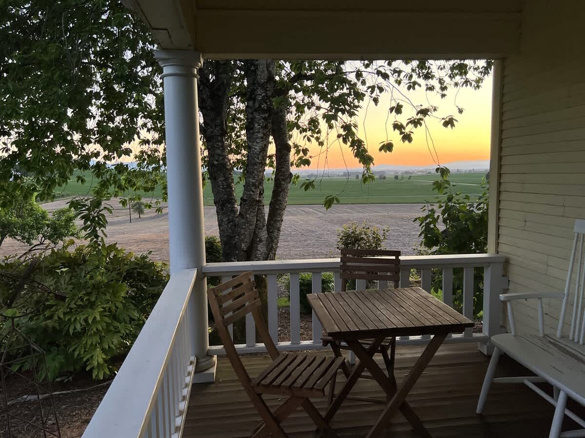 A cozy wooden porch features a small slatted table and two matching chairs, offering a serene view of a vast agricultural field under a vibrant sunset sky with hues of orange and yellow.