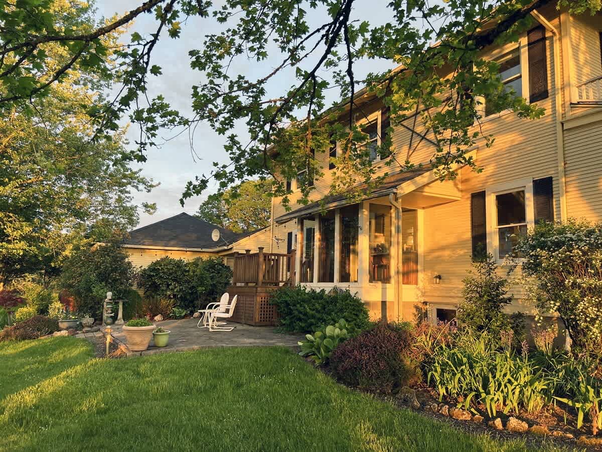 The side of the yellow farmhouse features a wooden deck with white rocking chairs and a paved patio below, surrounded by well-maintained landscaping and green lawn, bathed in the warm glow of late afternoon sun.