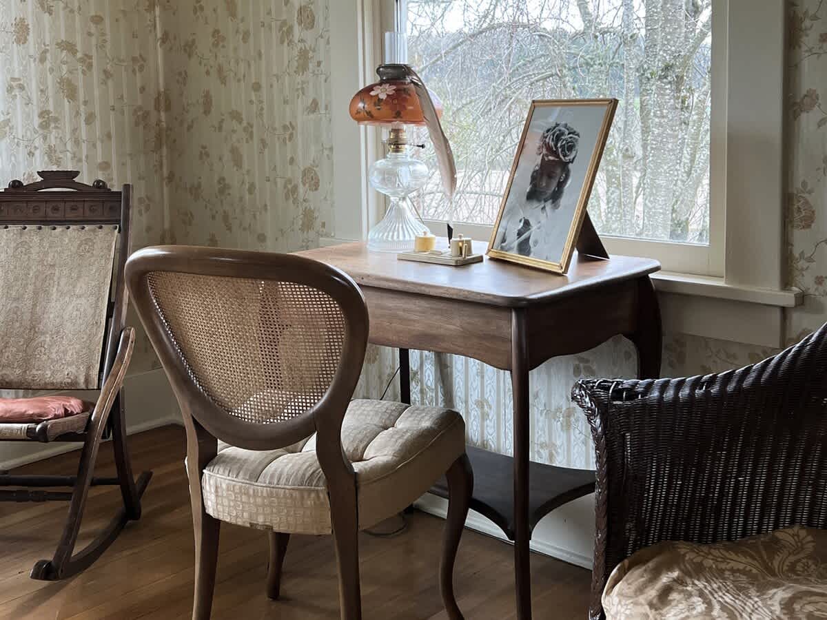 A cozy corner of a living space featuring a small wooden desk, a cane-back chair, and a vintage oil lamp, set against floral wallpaper with a window view of bare trees.