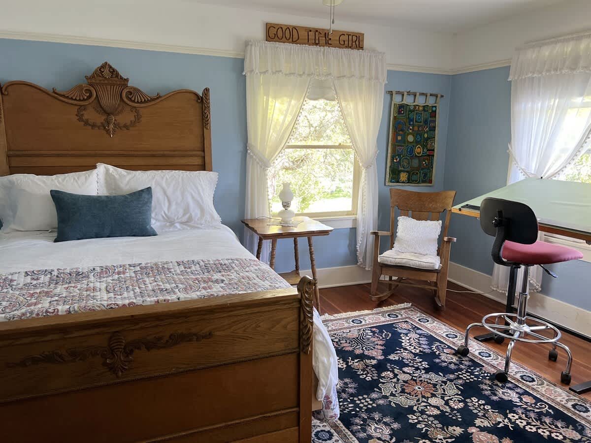 A charming bedroom with light blue walls, a carved wooden bed frame, a drafting table with a red chair, and a rocking chair, all on hardwood floors with a patterned rug.