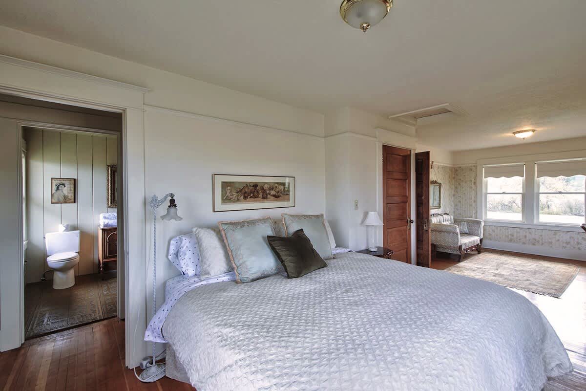 A spacious bedroom featuring a white quilted bed with light blue decorative pillows, a framed picture above the headboard, and hardwood floors, with an open doorway leading to a bathroom.